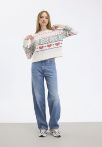 Knit sweater in cream with multicolored geometric and heart patterns. Paired with blue wide-leg jeans and patterned sneakers.