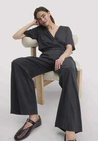 Dunkelgrauer Wickel-Jumpsuit mit kurzen Ärmeln, weitem Bein und taillierter Taille, kombiniert mit dunkelburgunderfarbenen, geschlossenen Schuhen. Texturierter Hintergrund.