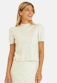 Kremowa bluzka o teksturze z wzorzystej tkaniny, z krótkimi bufkami i wysokim dekoltem, zdobiona pajęczynowatym wzorem i subtelnym połyskiem.