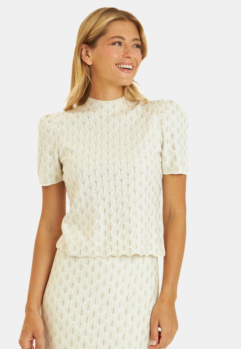 Kremowa bluzka o teksturze z wzorzystej tkaniny, z krótkimi bufkami i wysokim dekoltem, zdobiona pajęczynowatym wzorem i subtelnym połyskiem.