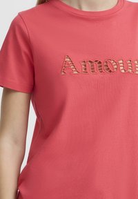 Korálově růžové tričko vyrobené z měkké tkaniny, s vyšitým textem "Amour" v vystřiženém stylu s pruhovanými detaily. Krátké rukávy a kulatý výstřih.