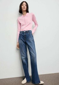 Helles rosa Langarm-Polohemd mit Knopfdetails, kombiniert mit hochgeschnittenen, ausgestellten blauen Jeans und beigen Slippern.
