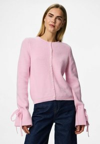 Sieviete, kas uzvelk gaiši rozā adītu kardiganu ar pērlītēm un saišķētiem piedurkņu galiem, ko apvieno ar tumši ziliem džinsiem uz baltas fona.