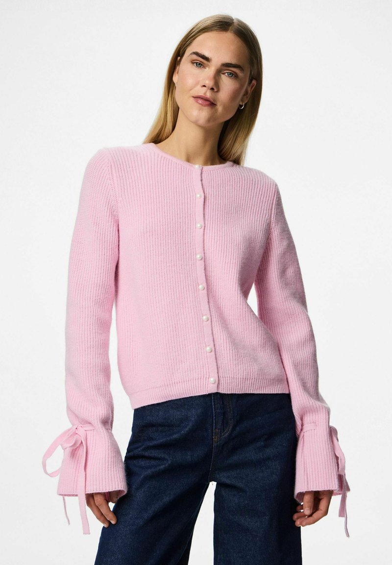 Sieviete, kas uzvelk gaiši rozā adītu kardiganu ar pērlītēm un saišķētiem piedurkņu galiem, ko apvieno ar tumši ziliem džinsiem uz baltas fona.