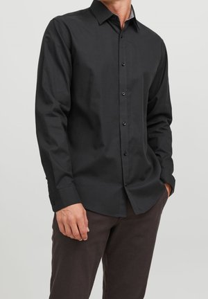 Chemise noire à manches longues avec col boutonné, fabriquée en tissu lisse, présentant une texture subtile et des boutons noirs. Portée avec un pantalon sombre.