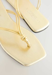 Guldiga platta sandaler med en slät metallisk yta, med tunna, korsade remmar och en fyrkantig tådesign. Elegant knoppspyndad detalj vid tån.