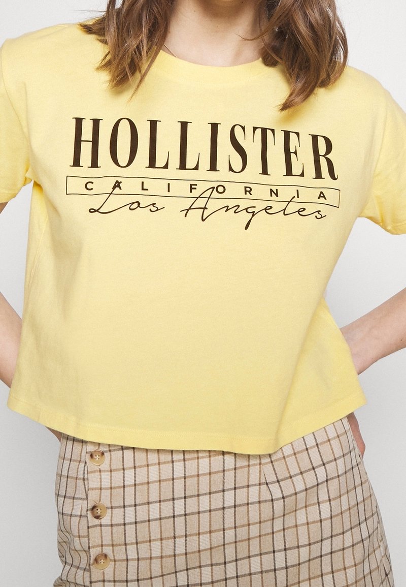 Žluté bavlněné zkrácené tričko s potiskem "HOLLISTER CALIFORNIA Los Angeles" v tmavě hnědé barvě. V kombinaci s béžovými kostkovanými kraťasy s vysokým pasem.