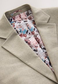 Blazer beige tissé avec un col à revers cranté révélant une doublure florale dans des tons de bleu, marron, rose et blanc à l'intérieur du col.