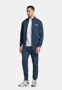 Tuta blu navy composto da giacca con zip e pantaloni, con logo bianco e strisce laterali a contrasto; abbinato a scarpe da ginnastica.