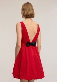 Punainen mekko, jossa syvä V-mallinen selkäosa, levenevä helma ja musta, paljetein koristeltu rusetti vyötäröllä. Valmistettu sileästä kankaasta, jolla on pehmeä tuntuma.