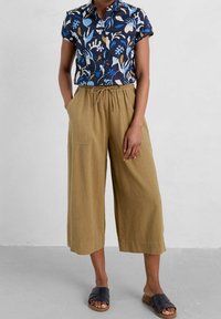 Person som bär en kortärmad marinblå blommig knappar-skjorta, lösa beige vida capribyxor och svarta slidesandaler, står med händerna i fickorna.