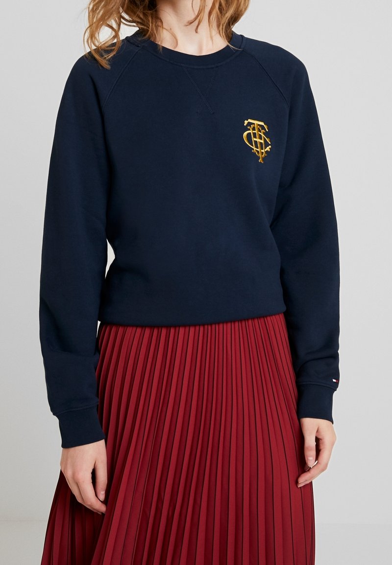 Marinblå sweatshirt med en guldbroderad logotyp på bröstet, parat med en röd pliserad kjol, med ribbade mudd och rundad hals.