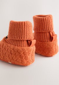 Chaussons pour bébé orange doux tricotés avec des poignets côtelés pliés et des motifs texturés, sur un fond neutre.