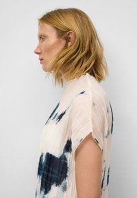 Vrouw met schouderlang blond haar, gekleed in een wit topje met korte mouwen en blauwe abstracte patronen, met haar gezicht naar links gericht tegen een eenvoudige witte achtergrond.