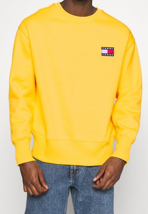 Mand iført en knaldgul Tommy Jeans sweatshirt og blå jeans, stående med hænderne afslappet ned langs siderne.