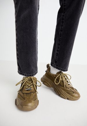 Osoba nosząca brązowe masywne sneakersy z siatkowymi i skórzanymi detalami, białe skarpetki oraz czarne jeansy denimowe stojąca na białej powierzchni.