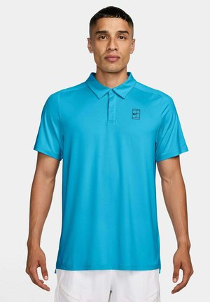 Man draagt een felblauw poloshirt met korte mouwen en een klein zwart logo op de linkerborst, gecombineerd met witte shorts.