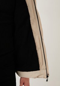 Giacca a vento beige con interno nero peloso, dotata di apertura con zip e testi in contrasto sulle maniche.