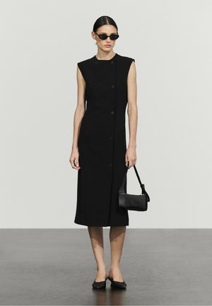 Femme debout portant une robe midi noire sans manches boutonnée, des ballerines noires, des lunettes de soleil et tenant un petit sac à main noir.