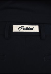 Gros plan sur la ceinture d’un pantalon noir avec une étiquette rectangulaire en tissu blanc brodée du mot « Prohibited » en cursive noire.