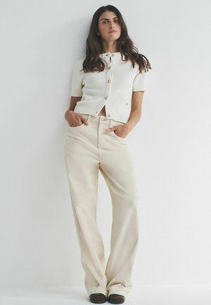 Cremefarbener Kurzarm-Cardigan mit strukturiertem Muster und goldenen Knöpfen, kombiniert mit beigefarbenen Hosen mit weitem Bein und dunklen Schuhen.