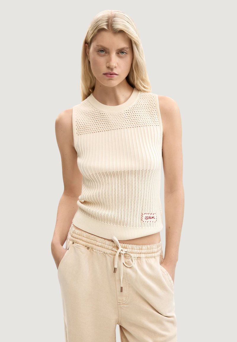 Jeune femme aux longs cheveux blonds portant un haut en tricot crème sans manches et un pantalon beige à cordon, les mains dans les poches.