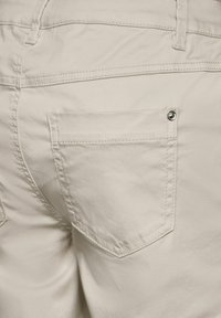 Beige cotton pants featuring a rear pocket with a silver snap button. Smooth texture and visible stitching along the waistband and pocket edges.
