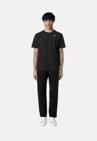 T-shirt nera in cotone con maniche corte, colletto tondo e un piccolo logo sul petto, abbinata a pantaloni neri slim-fit e sneakers bianche.