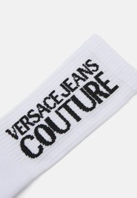Bela rebrasta nogavica s črnimi napisi "VERSACE JEANS COUTURE". Material se zdi mehak, z teksturirano površino in izrazito tipografijo.