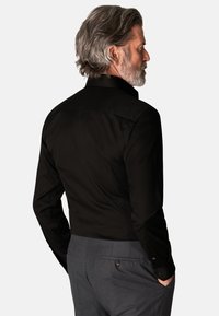 Svart langermet skjorte med strukturert krage, glatt tekstur og tettsittende design, kombinert med grå bukser med en knappet baklomme.