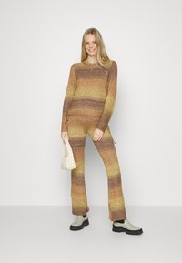 Conjunto de suéter e calças tricotadas amarelo e castanho listradas, com um corte descontraído, acabamento texturizado e fendas laterais. Os acessórios incluem uma mala creme.