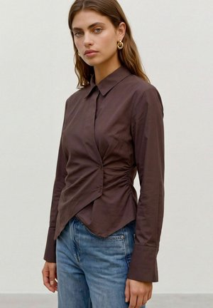 Modèle portant un chemisier ajusté marron foncé, boutonné sur le devant et froncé à la taille, un jean bleu et des créoles dorées.