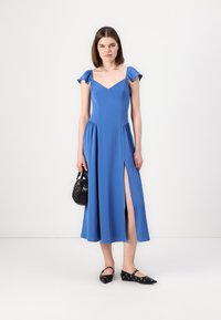 Robe bleue, longueur genoux, avec un décolleté en V et des manches courtes à volants. Présente une fente sur le côté et une jupe fluide. Sac à main noir et chaussures plates.