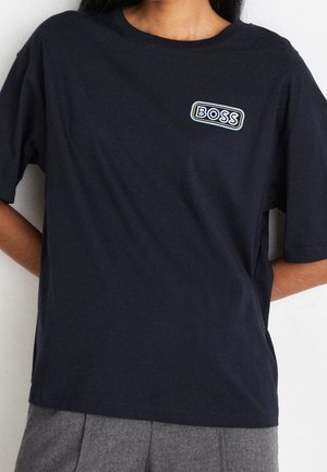 Persoon draagt een zwart T-shirt met een klein "BOSS"-logo op de borst, gecombineerd met een grijze broek, tegen een effen lichte achtergrond.