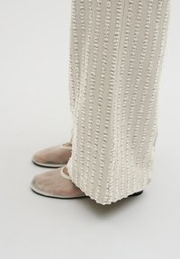 Translucente beige Sandalen mit offenem Zehendesign, getragen mit cremefarbenen, strukturierten Hosen, die vertikale Streifen und erhabene Akzente aufweisen.