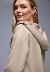 Beige hættetrøje med et mønstret gråt hættestof, der har lynlås og glat tekstur. Casual pasform med sænkede skuldre.