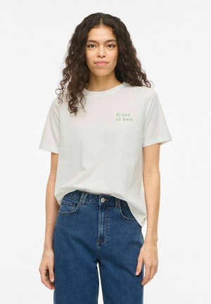 Femme aux cheveux bouclés portant un t-shirt blanc avec l'inscription « C’est si bon » et un jean bleu taille haute, debout sur un fond blanc.
