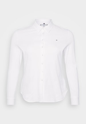 Hvit skjorte med lange ermer og krage med knapper, med et brystlogo, en glatt tekstur og bølget nederkant. Knapper er jevnt plassert.