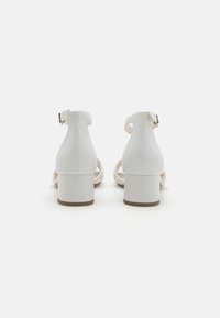 White heeled sandals made of smooth leather, featuring an open back, buckle strap closure, and block heels. Sole is light brown.