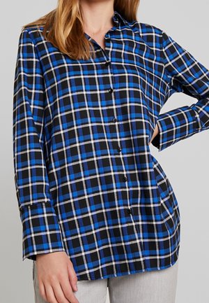 Frau, die ein blau-schwarz-weiß kariertes Hemd mit langen Ärmeln trägt und mit einer Hand in der Hüfte steht.