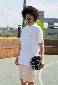 Hvid kortærmet skjorte med et pink og blåt Nike-logo, parret med lyserøde shorts med sorte detaljer, mens personen holder en sort basketball.