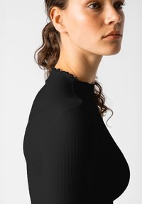 Schwarzes geripptes Oberteil mit einem hohen, gewellten Ausschnitt. Der Stoff wirkt glatt mit vertikalen Rillen. Das Haar des Models ist leicht gewellt.