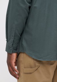 Camisa de manga larga verde oscuro con puños abotonados, combinada con pantalones color crema que tienen bolsillos laterales. Textura suave, corte relajado, diseño casual.