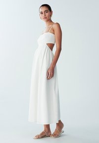 Valge linane maxi-kleit, millel on kujundatud korsett, spagetitikujulised rihmad, külgavad lõiked ja voogav seelik. Kandatud koos beežide rihmikute sandaalidega.