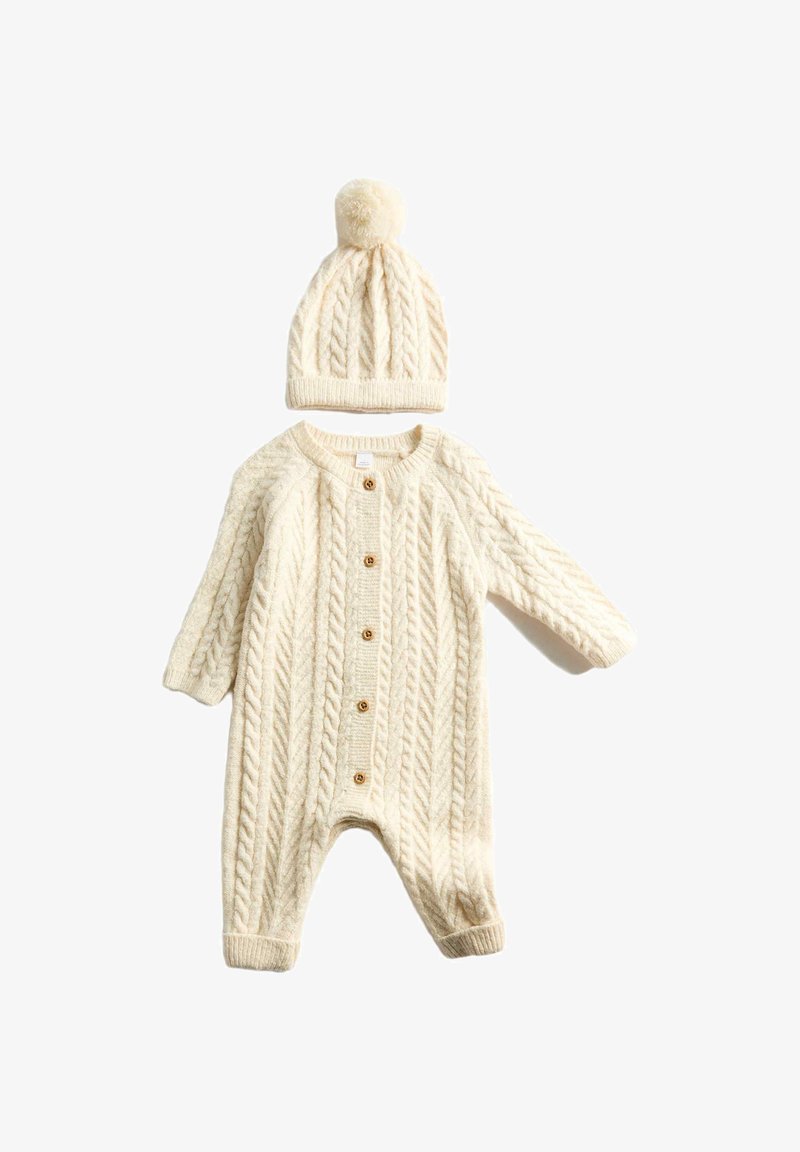Krémový pletený overal s kabelovým vzorem, kulatým výstřihem, dlouhými rukávy a dřevěnými knoflíky. Součástí je také čepice se střapcem.