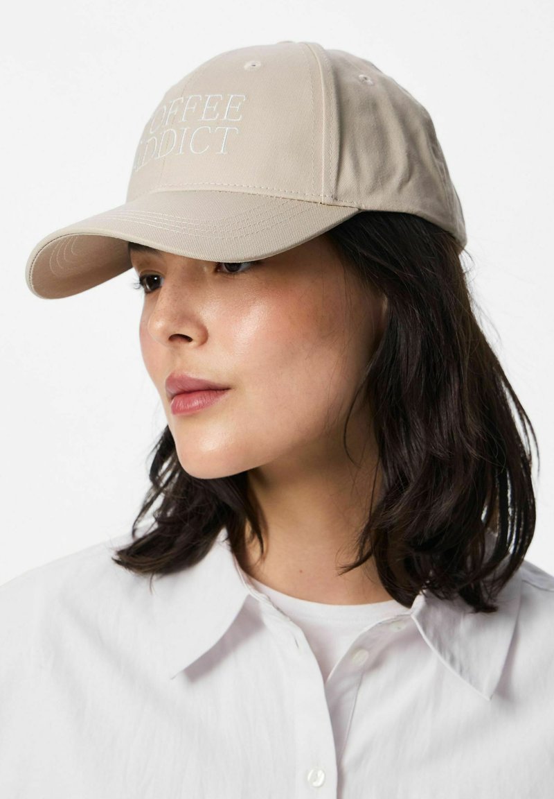 Jeune femme aux cheveux foncés portant une casquette beige avec l'inscription « COFFEE ADDICT » et une chemise blanche à col, regardant légèrement sur le côté.