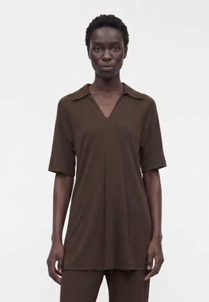 Femme noire portant un t-shirt marron ample avec col en V, col et manches courtes, debout devant un fond blanc uni.