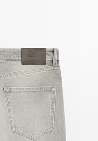 Jeans grigi chiari con tessuto testurizzato, dotati di un'etichetta in pelle marrone sulla vita posteriore e una singola tasca posteriore.