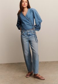 Denim wrap top s puff rukavima, uparen s svijetlosivim širokim trapericama koje imaju detalj od lanca. Smeđe ravne cipele upotpunjuju odjevnu kombinaciju.