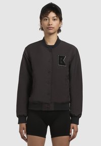 Must jakk musta värvi, ümmarguse kaelusega, nööpidega esiosas ja sooniknæppudega. Vasakul rinnal on tekstuuriga lapiga, millel on 'K'. Minimalistlik disain.